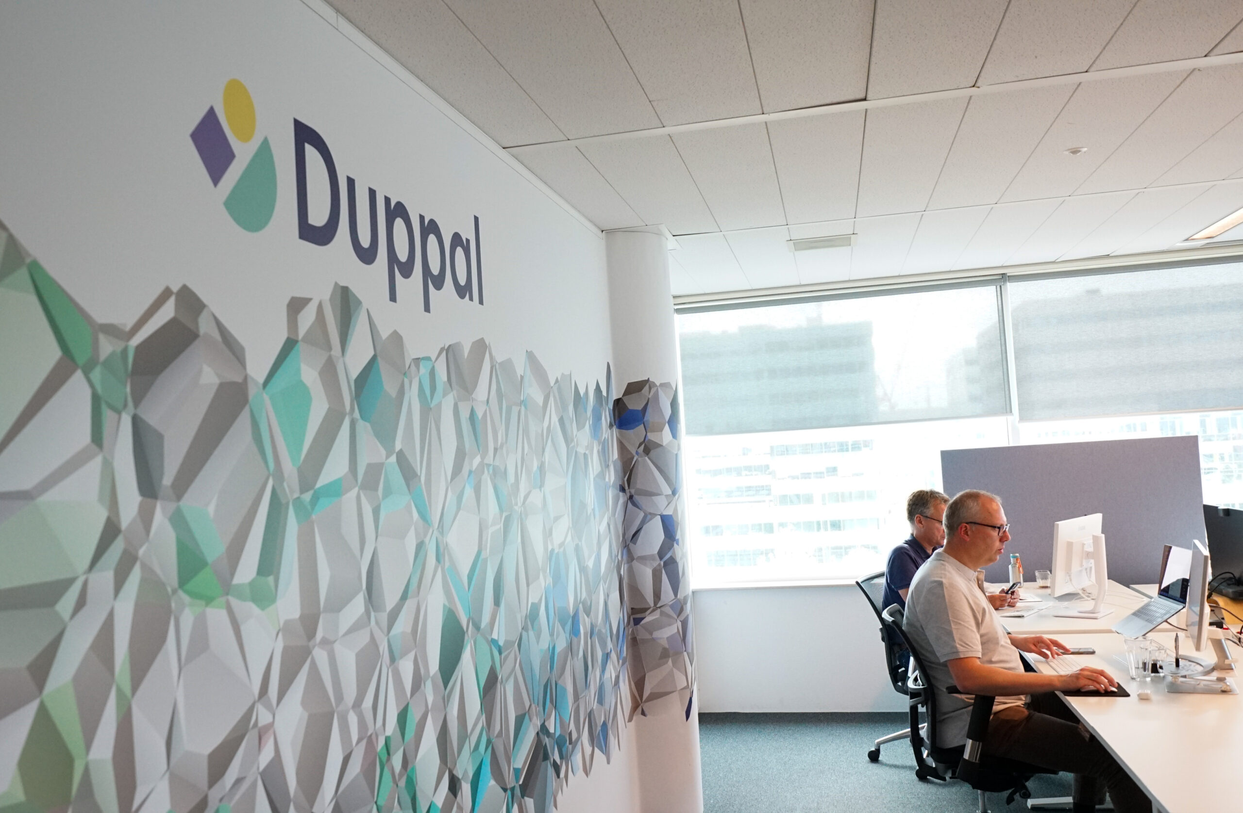 Het Duppal-kantoor in Amsterdam, met Duppal-logo en brand-pattern op de muur en twee collega's aan het werk aan een bureau.